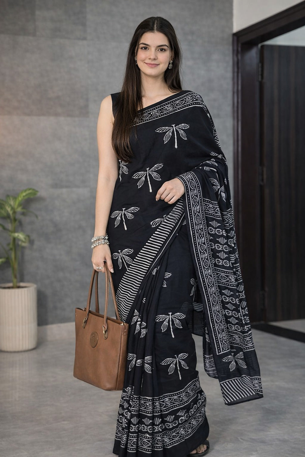 Woman wearing a black saree with white patterns, holding a brown handbag indoors.